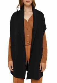 Zwarte mouwloze cardigan met een sjawl kraag en twee voorzakken, gelaagd over een gestructureerde bruine knooptrui.