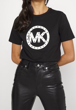 Czarny bawełniany t-shirt z dużym białym okrągłym logo z napisem "MK" w centrum, otoczonym srebrnymi ćwiekami. Połączony z czarnymi skórzanymi spodniami.