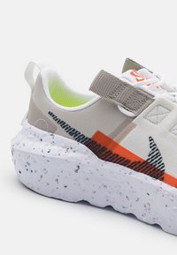 Gros plan d'une basket blanche et grise avec une semelle texturée, des accents orange et noirs, une bride Velcro et une languette au talon.