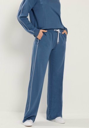 Person in blauer Jogginghose mit weißen Seitenstreifen und passendem Sweatshirt, Hände in den Taschen, steht auf weißem Boden.