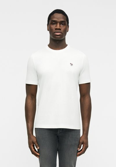 Jeune homme debout, face à la caméra, portant un t-shirt blanc uni à manches courtes avec un petit logo zèbre multicolore sur la poitrine.