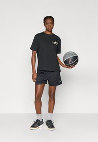 Μαύρο βαμβακερό μπλουζάκι με γραφικό «GENERATION», συνδυασμένο με μαύρες κοντές μπλούζες, γκρι κάλτσες και μαύρα αθλητικά παπούτσια, κρατώντας μια μπάλα μπάσκετ.