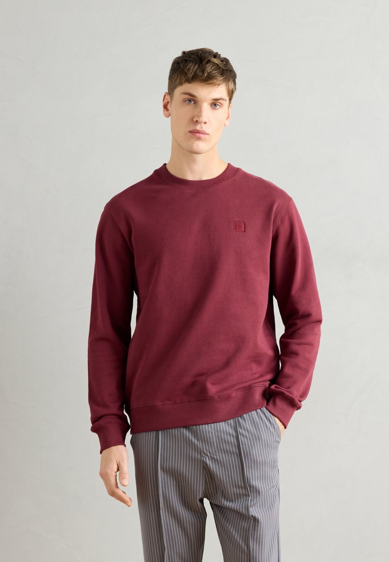 Sudadera burdeos con mangas largas y puños acanalados. Presenta un logo cuadrado en el pecho y un cuello redondo. Combinada con pantalones a rayas.