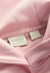 Jasnoróżowy bawełniany odzież zbliżenie na kołnierzyk. Metki z napisem "BAKER by TED BAKER" oraz "MADE IN BANGLADESH" w neutralnych odcieniach.