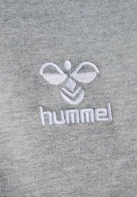 Sivi materijal s glatkom teksturom sa bijelim izvezenim logotipom stilizirane figure iznad natpisa "hummel."