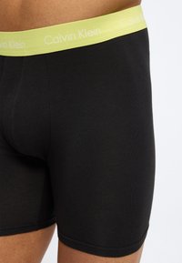 Μαύρα ανδρικά μποξεράκια με φάρδος μέσης σε λαχανί χρώμα και λευκά λογότυπα Calvin Klein, φορεμένα από άτομο μπροστά σε μονόχρωμο φόντο.