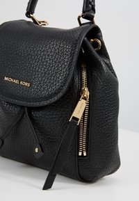 Sac à main Michael Kors en cuir noir texturé avec fermeture éclair dorée et quincaillerie, fermeture à rabat et poignée supérieure sur une surface unie.