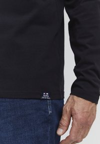 Fekete, hosszú ujjú póló, puha pamutból készült, bordázott gallérral. Az alján látható a "FQ1924 ORIGINAL" felirat. Kék farmernadrág részben látható.