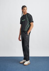 Černé sportovní tričko s zelenými akcenty a logem, v kombinaci s černými sportovními kalhotami a bílými teniskami. Hladký materiál a volný střih.