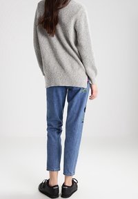 Grau strukturierter Pullover, blaue Jeans mit floralem Stickmuster und schwarze Sneakers. Das Outfit hat eine lockere Passform und ein lässiges Design.