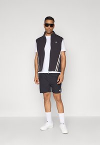 Träningsväst, svart tyg med crèmefärgade ränder, vit t-shirt, svarta shorts och vita sneakers. Stora svarta solglasögon. Enkelt mönster.