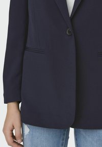 Jūras spēku žakete ar vienu pogu, divām priekšējām kabatām un dekoratīvo šuvju apdari. Valkāta virs gaišas krāsas krekla, kombinēta ar nodilušām džinsiem.