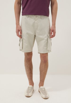 Mann trägt beige Cargo-Shorts, weiße Schnürsneaker und ein dunkelrotes T-Shirt, steht vor einem einfarbigen Hintergrund.