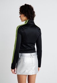 Jachetă neagră cu mâneci lungi, guler înalt și dungi neon verzi, asortată cu o fustă mini strălucitoare, argintie, cu un model texturat.