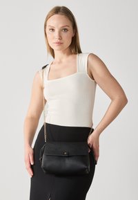 Borsa a tracolla in pelle nera con catena, caratterizzata da un pattino ripiegato e dettagli in oro. Il modello indossa un top aderente crema e una gonna nera.