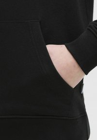 La felpa nera presenta una tasca a marsupio con apertura visibile, polsini a coste e una texture morbida, mostrando un design elegante e minimalista.