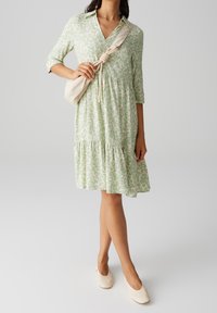 Robe vert clair avec un décolleté en V, des manches trois-quarts, une taille froncée et une jupe à volants, ornée d'un motif floral. Associée à un sac beige.