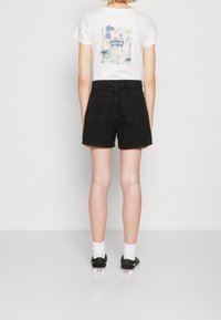 Pantalones cortos de mezclilla negra con corte recto, dos bolsillos traseros y dobladillos enrollados, combinados con una camiseta blanca con un gráfico floral colorido.
