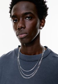 Grå sweatshirt med ribbet krave, lagdelte sølvkæder og en rund ørestik. Glat hudstruktur og tætklippet hår.