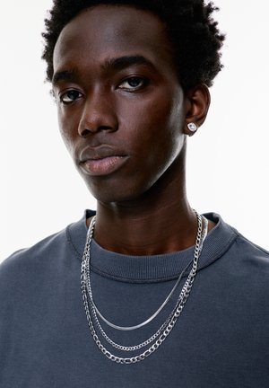 Sweatshirt gris avec col côtelé, chaînes en argent superposées et une boucle d'oreille ronde. Texture de peau lisse et cheveux très courts.