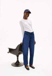 Blauwe denim high-waisted jeans, witte textuur lange mouwen top, marineblauwe baseball cap, zwarte platte schoenen met decoratieve hardware, zwarte stoel.