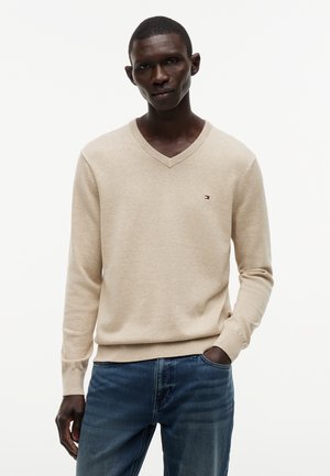 Άνδρας που φοράει μπεζ πουλόβερ με V λαιμόκοψη και μικρό λογότυπο στο στήθος, καθώς και μπλε τζιν, στέκεται με ένα χέρι στην τσέπη μπροστά σε απλό φόντο.