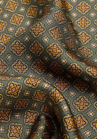 Tejido verde con patrones florales ornamentales repetidos en amarillo, marrón y rojo, mostrando pliegues y una textura en relieve.