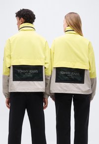 Δύο άτομα φορούν κίτρινες και γκρι μπουφάν Tommy Jeans με μαύρες τσέπες από δίχτυ στην πλάτη, στέκονται το ένα δίπλα στο άλλο με την πλάτη προς το μέρος μας.