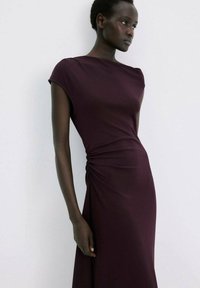 Vrouw met een donkere huid die een mouwloze, nauwsluitende donkerpaarse jurk draagt met geplooide details aan de zijkant, staand tegen een witte achtergrond.