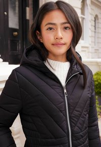 Chaqueta de plumas negra con capucha, que presenta un patrón de acolchado diagonal, cremallera plateada y un suéter blanco acogedor debajo.