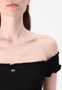 Top noir décolleté en tissu texturé et froncé. Présente des bords volants et un petit logo brodé en rouge et blanc.