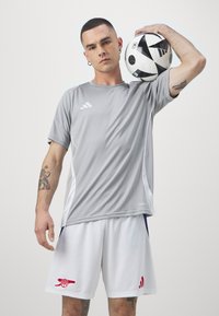 Camiseta atlética gris con acentos blancos, combinada con pantalones cortos blancos que tienen un logo rojo. La persona sostiene un balón de fútbol en blanco y negro.