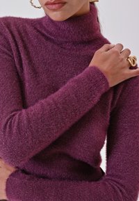 Donna che indossa un maglione a collo alto viola morbido, si tiene la spalla, indossa un anello d'oro intrecciato e un grande orecchino a cerchio d'oro.