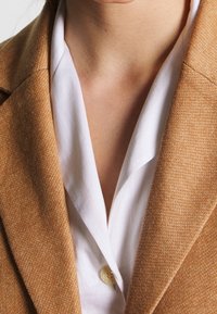 Primer plano de una persona que lleva puesta una chaqueta marrón claro sobre una camisa blanca con cuello y el botón superior desabrochado.