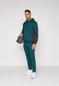 Conjunto de hoodie e calças azul-petróleo com detalhes pretos; feito de tecido macio; inclui um bolso frontal; combinado com uma mala preta e ténis brancos.