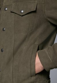 Olive green suede jacket with black snap buttons, two chest pockets, a smooth texture, and a side pocket. Tailored design with a fitted shape.