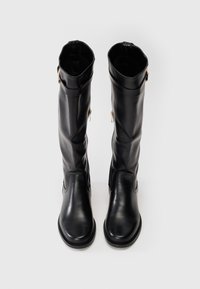 Um par de botas de couro pretas até ao joelho com fechos laterais e tiras de fivela sobre um fundo neutro.