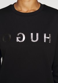 Schwarzes Langarm-T-Shirt aus weichem Stoff mit einem glänzenden silbernen Schriftzug "HUGO" in Blockbuchstaben.