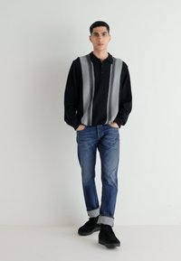 Polo negro de mangas largas con rayas verticales grises, combinado con vaqueros azules remangados en los puños y zapatos negros. Diseño sencillo y moderno.