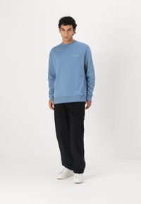 Light blue sweatshirt made of cotton, featuring a crew neck and understated text on the chest. Paired with black wide-leg trousers and white sneakers.