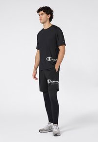 Joven de pie vistiendo una camiseta atlética negra de Champion, pantalones cortos, mallas negras y zapatillas grises de Champion, de perfil hacia la izquierda sobre un fondo blanco.