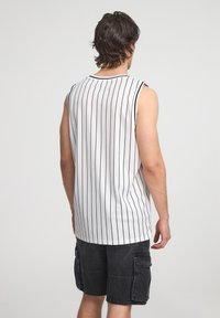 Camisetta bianca senza maniche con strisce verticali nere, caratterizzata da un collo rotondo. Indossata con pantaloni cargo neri. Design semplice, tessuto leggero.