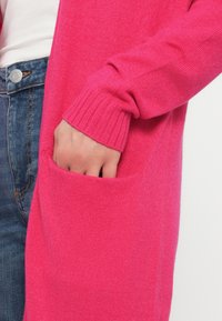Μακρύ, ζωντανό ροζ ζακέτα από απαλό πλεκτό ύφασμα. Διαθέτει ριμπ μανσέτες και μια μπροστινή τσέπη, με άνετη εφαρμογή.