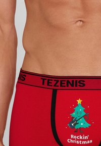 Czerwone bawełniane bokserki z czarnym elastycznym paskiem w logo "TEZENIS" oraz świąteczną grafiką w formie rysunkowej choinki.