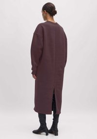 Lang, oversized jurk van donkerpaarse stof met een strak ontwerp, wijde mouwen en een split aan de achterkant, gecombineerd met zwarte knielange laarzen.