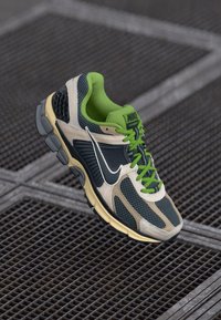 Nike running shoe featuring a mesh upper in gray and beige, green laces, and a black swoosh. Textured sole for traction.