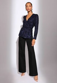 Navy-blaues Top mit Pailletten, Wickeldesign und V-Ausschnitt, kombiniert mit hochtaillierten, schwarzen, weiten Hosen und klaren Absatzsandalen.