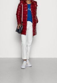 Red, puffy coat with a hood and glossy finish, worn over a blue t-shirt, paired with white trousers and translucent-sole sneakers.