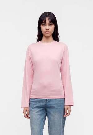 Femme aux cheveux raides et foncés portant une chemise rose clair à manches longues et un jean bleu clair, debout devant un fond blanc uni.