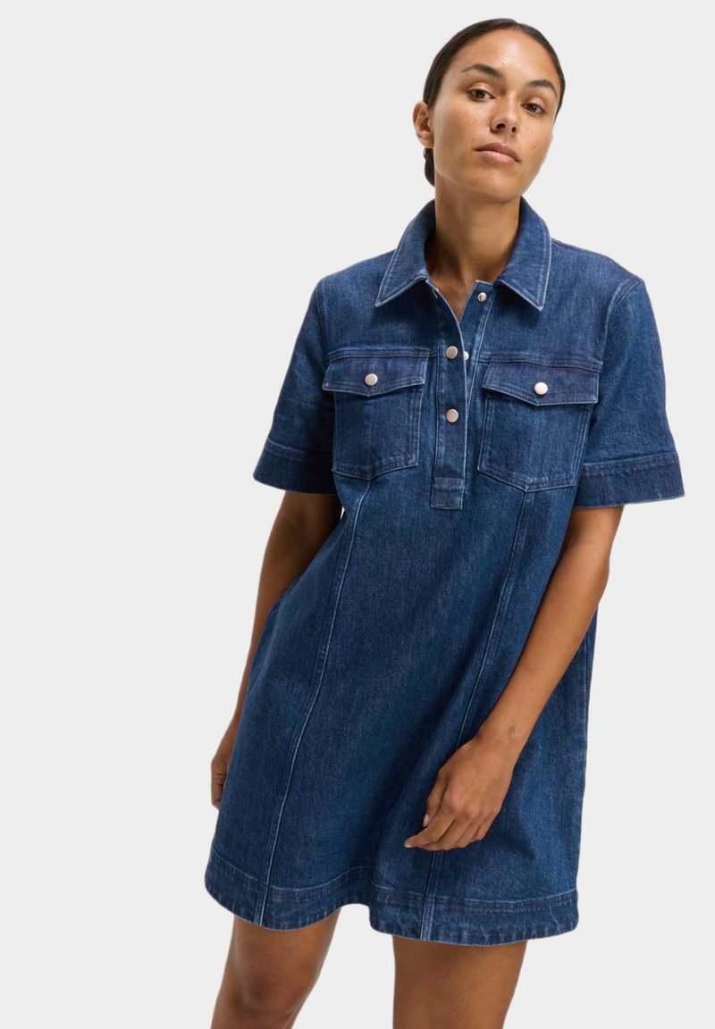 Vestido de camisa de denim en azul oscuro, con cuello, mangas cortas, dos bolsillos en el pecho con solapas y cierre de botones. Textura suave.
